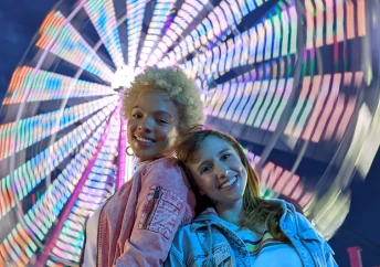 Two young women smiling and posing in front of a ferris wheel at night.